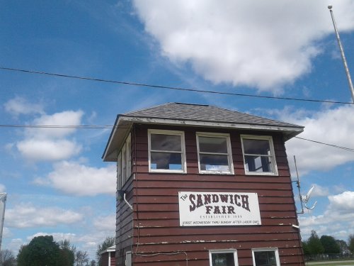 A red wooden building with a sign reading "The Sandwich Fair" against a blue sky with fluffy clouds.