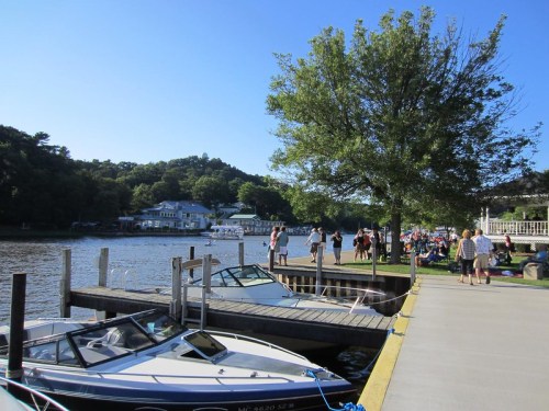 A sunny lakeside scene with boats docked, people walking along the shore, and trees in the background.