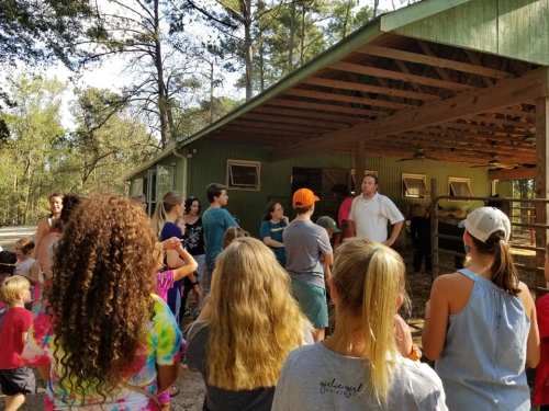 A group of people, including children, gather outside a barn for a presentation in a wooded area.
