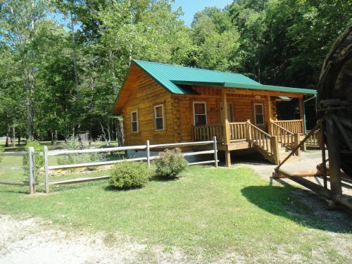 A small wooden cabin with a green roof, surrounded by trees and a grassy area, featuring a front porch and wooden railing.