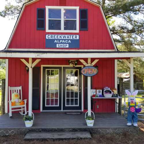 A red barn-style shop with a sign reading "Creekwater Alpaca Shop," featuring colorful decorations and an open sign.