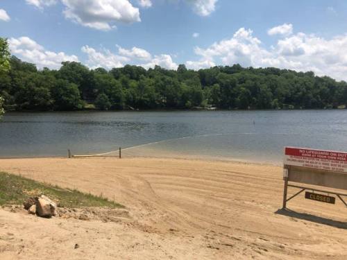 A sandy beach area by a calm lake, surrounded by green trees, with a "Closed" sign visible. Fluffy clouds in the sky.