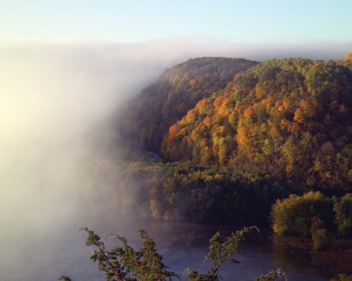 A misty landscape featuring a colorful forested hill overlooking a river, with fog enveloping the scene.