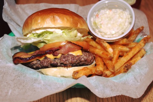 A juicy burger with lettuce, tomato, cheese, and bacon, served with fries and a side of coleslaw in a basket.