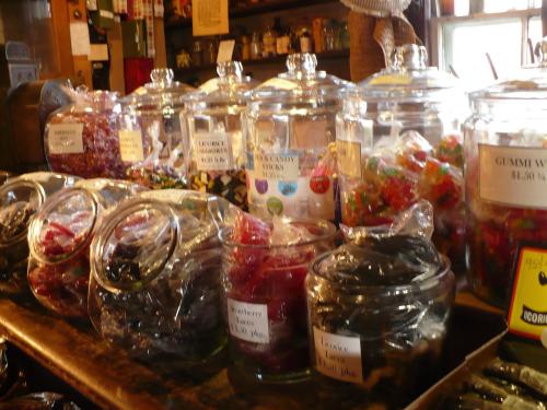 A variety of colorful candies in glass jars on a wooden counter, with labels indicating different types and prices.