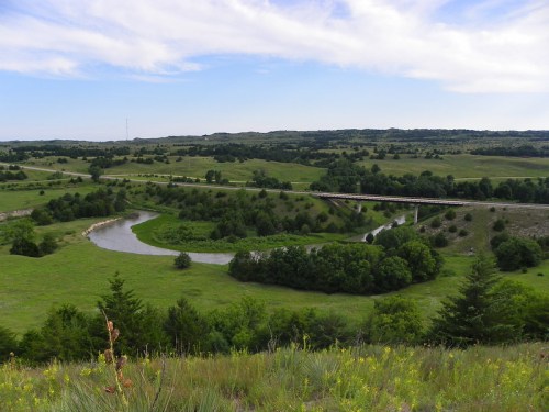 A scenic view of a winding river surrounded by lush green hills and trees under a blue sky with scattered clouds.