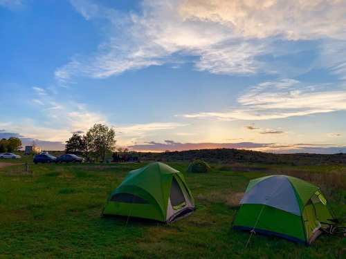 Two green tents set up on grassy land under a colorful sunset sky, with distant hills and cars in the background.