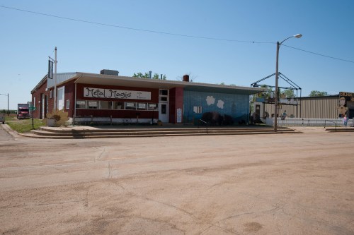 A small, colorful building with a mural, labeled "Metal Magic," on a quiet street under a clear blue sky.