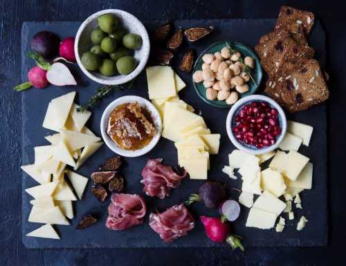 A charcuterie board featuring assorted cheeses, olives, nuts, figs, pomegranate, radishes, and crackers on a dark slate.