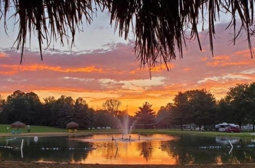 A serene pond at sunset, with a fountain and silhouettes of trees against a colorful sky.