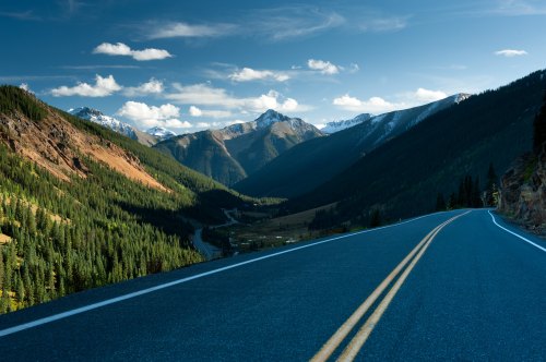 A winding road through a mountainous landscape, surrounded by lush forests and snow-capped peaks under a blue sky.
