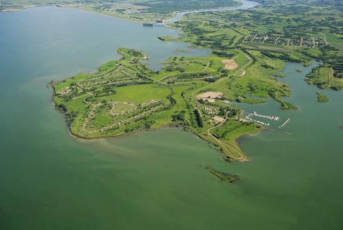 Aerial view of a green island surrounded by water, with a marina and land in the background.
