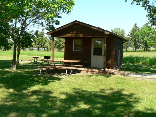 A small wooden cabin surrounded by trees and picnic tables on a grassy area.