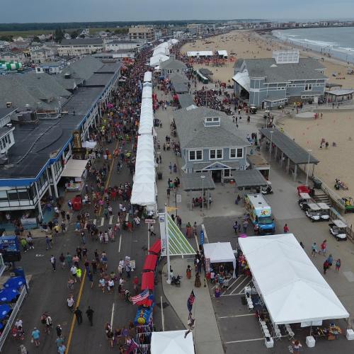 Aerial view of a crowded beach boardwalk with tents, shops, and a sandy beach in the background.