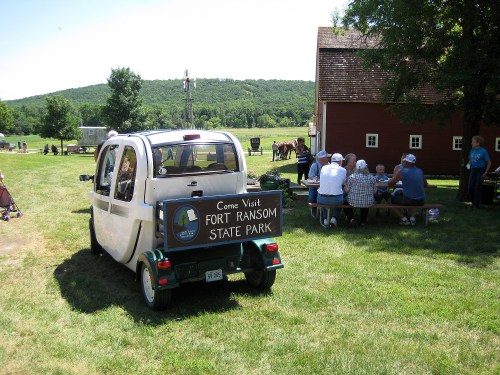 A small electric vehicle parked on grass, with a sign promoting Fort Ransom State Park and people gathered at a picnic table.