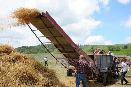 Farmers operate a vintage hay baler, unloading straw onto a large pile under a blue sky with rolling green hills.