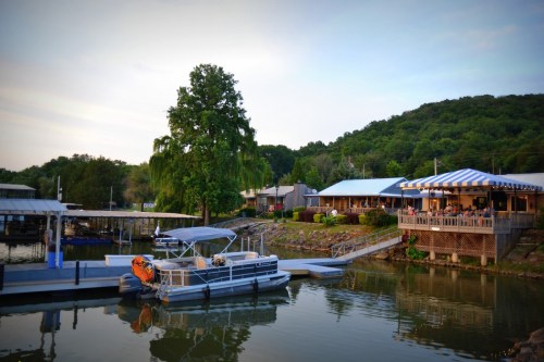 A serene lakeside scene featuring a docked boat, a restaurant, and lush green hills in the background.