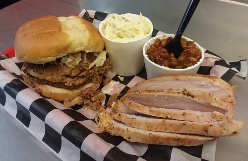 A pulled pork sandwich with coleslaw, served with sides of potato salad and baked beans on a checkered paper.