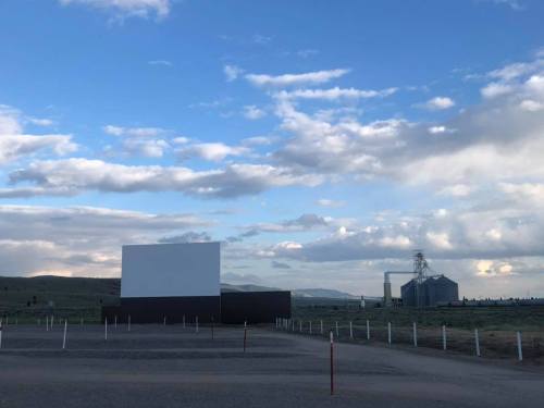 A large blank movie screen stands in an open field under a cloudy sky, with a building and silos in the background.
