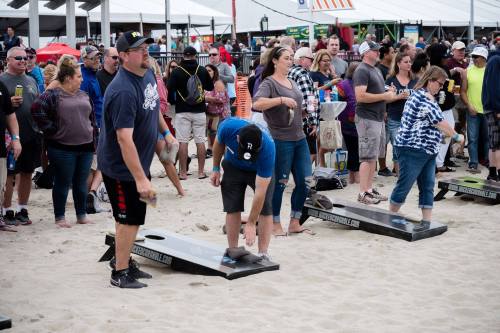 A crowd watches as participants play cornhole on the beach during a lively outdoor event.