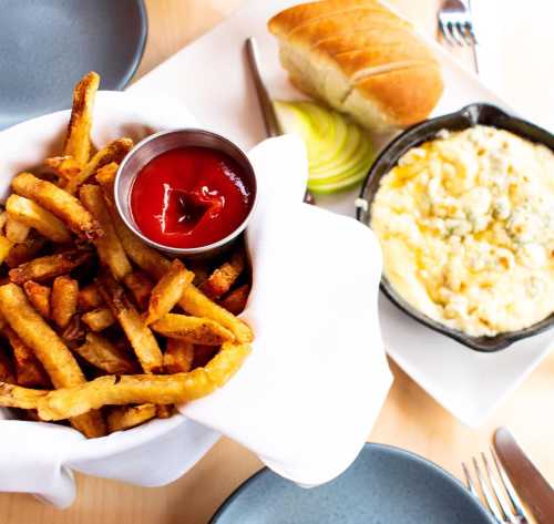 A bowl of golden fries with ketchup, a side of baked dish, and a roll on a table set with plates and utensils.