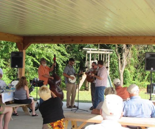 A bluegrass band performs under a pavilion while an audience enjoys the music. Trees are visible in the background.