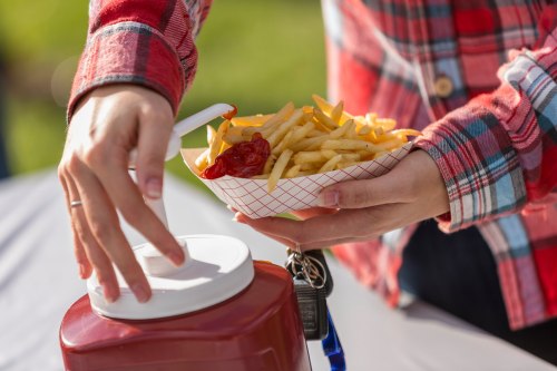 A person in a plaid shirt holds a tray of fries while reaching for a ketchup dispenser.