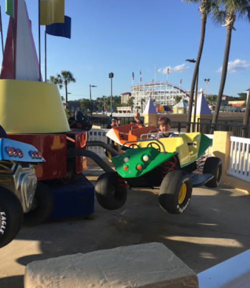 Children enjoy colorful ride-on cars at an amusement park, with a roller coaster visible in the background.