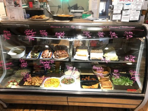 Display case filled with various food items, including meats, salads, and sides, with handwritten menu items and prices.