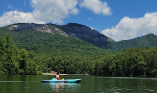 A person kayaking on a calm lake surrounded by lush green mountains and a blue sky with fluffy clouds.