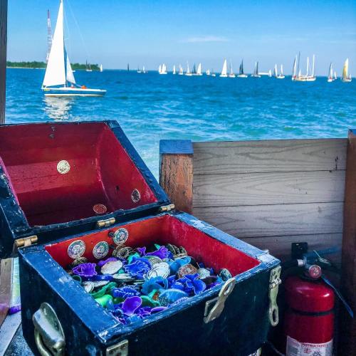 A treasure chest filled with colorful coins sits on a dock, with sailboats in the background and a clear blue sky.