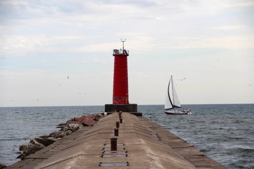 A red lighthouse stands on a pier, with a sailboat passing by on a calm sea under a cloudy sky.