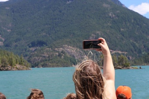 A person with long hair holds a phone up to capture a scenic view of mountains and a turquoise lake.