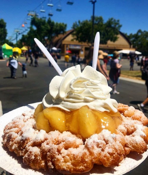A plate of funnel cake topped with whipped cream and apple filling, with a fair in the background.