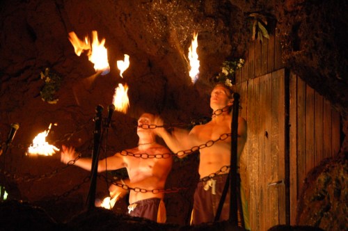 Two shirtless performers juggling flaming torches in a cave setting, surrounded by chains and dim lighting.