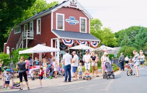 A lively scene outside a red general store, with families, children, and vendors enjoying a community event.