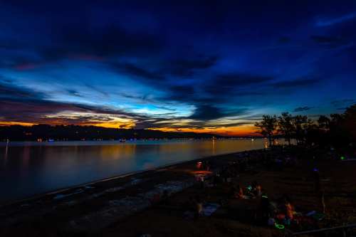A serene lakeside view at dusk, with vibrant clouds reflecting on the water and distant lights twinkling along the shore.
