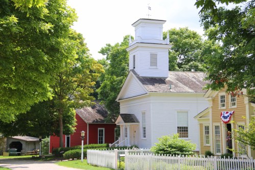 A quaint village scene featuring a white church, red building, and lush greenery under a bright blue sky.