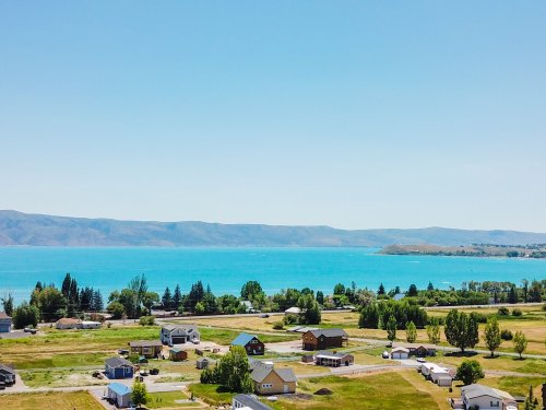 A scenic view of a lake surrounded by green fields and houses, with mountains in the background under a clear blue sky.