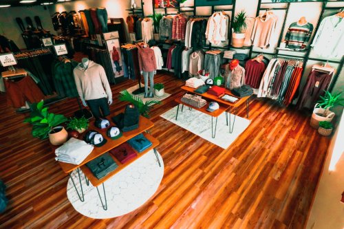 A clothing store interior featuring neatly arranged apparel on racks and tables, with wooden floors and plants.