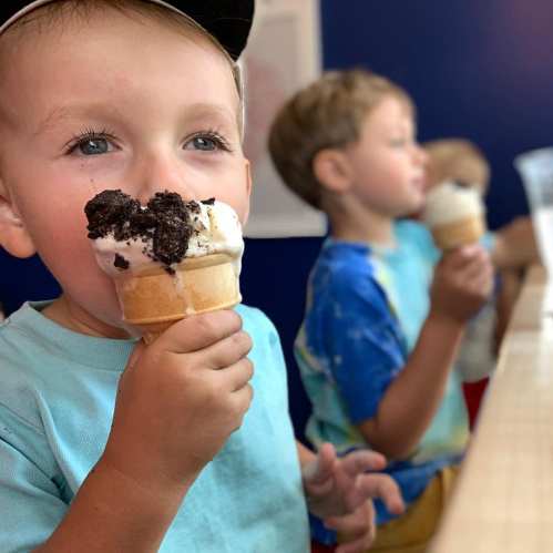 A young child with a blue cap enjoys an ice cream cone, with other children in the background also holding ice cream.