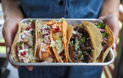 A person holding a tray with four colorful tacos filled with various ingredients.
