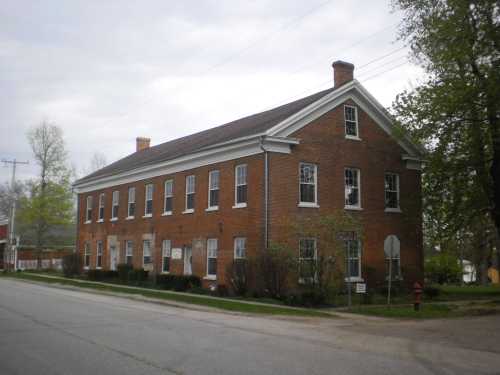 A two-story brick building with multiple windows, set on a quiet street lined with trees.