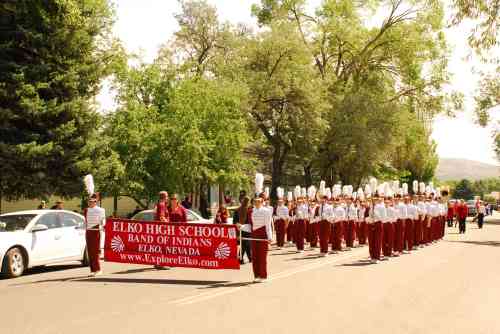 A marching band in red and white uniforms parades down a tree-lined street, holding a banner for Elko High School.