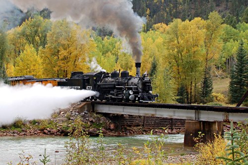 A vintage steam train crosses a bridge over a river, surrounded by vibrant autumn foliage.