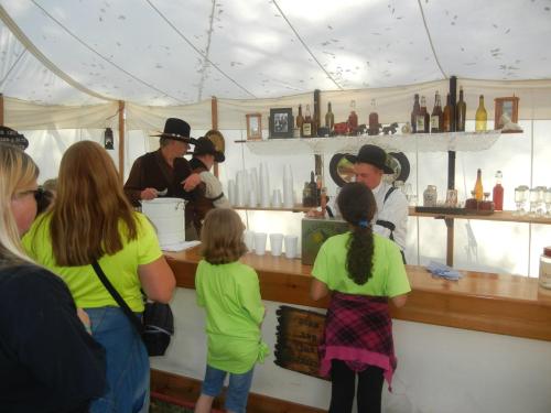 A group of people at a bar serving drinks under a tent, with a bartender and customers in period costumes.