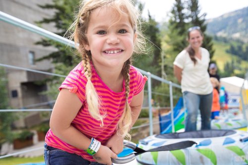 A smiling girl with braids holds a handle on a colorful inflatable slide, with adults and trees in the background.