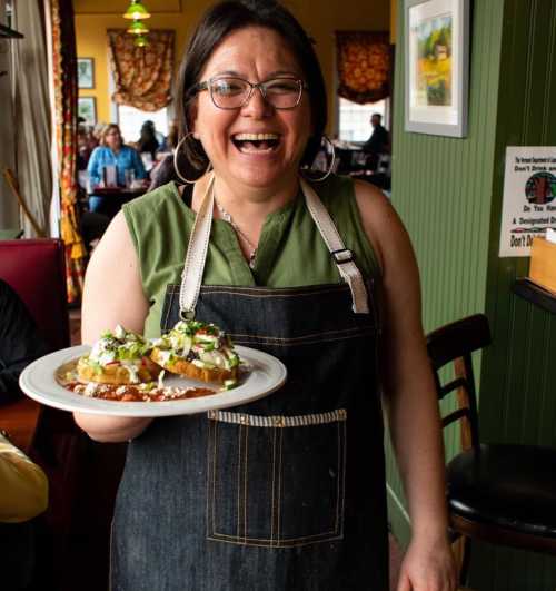 A smiling woman in an apron holds a plate of food in a cozy restaurant setting.