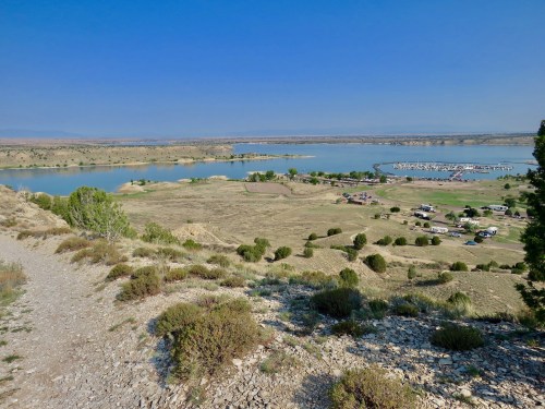 A scenic view of a lake surrounded by hills and greenery, with boats docked and a clear blue sky above.