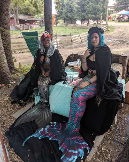 Two mermaids in colorful tails sit on a couch, surrounded by nature, with snacks and drinks nearby.